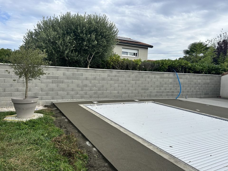 Chape béton lissée fraîchement coulée en ceinture autour d'une piscine à volet roulant, avant la pose du bois